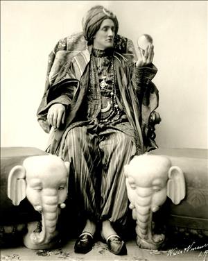 A person is seated between two ottomans decorated with ivory elephant heads. The person wears loose striped pants and robes and pointy shoes. They have a turban on their head and are holding up a glass ball that they gaze into. 